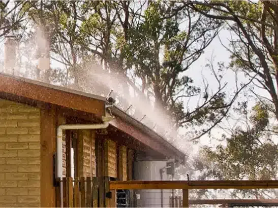 Sprinklers mist water over a house roof, likely to protect from fire, with trees in background.
