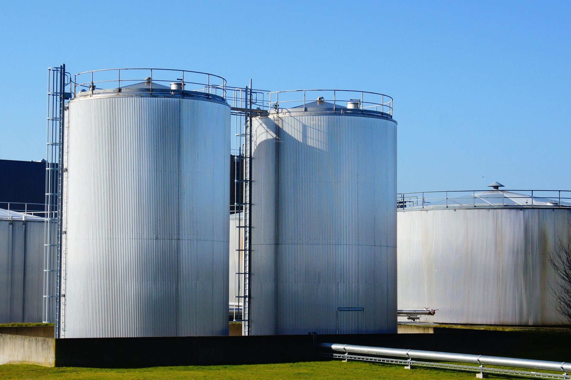 Three large white industrial storage tanks on a grassy site under a clear blue sky