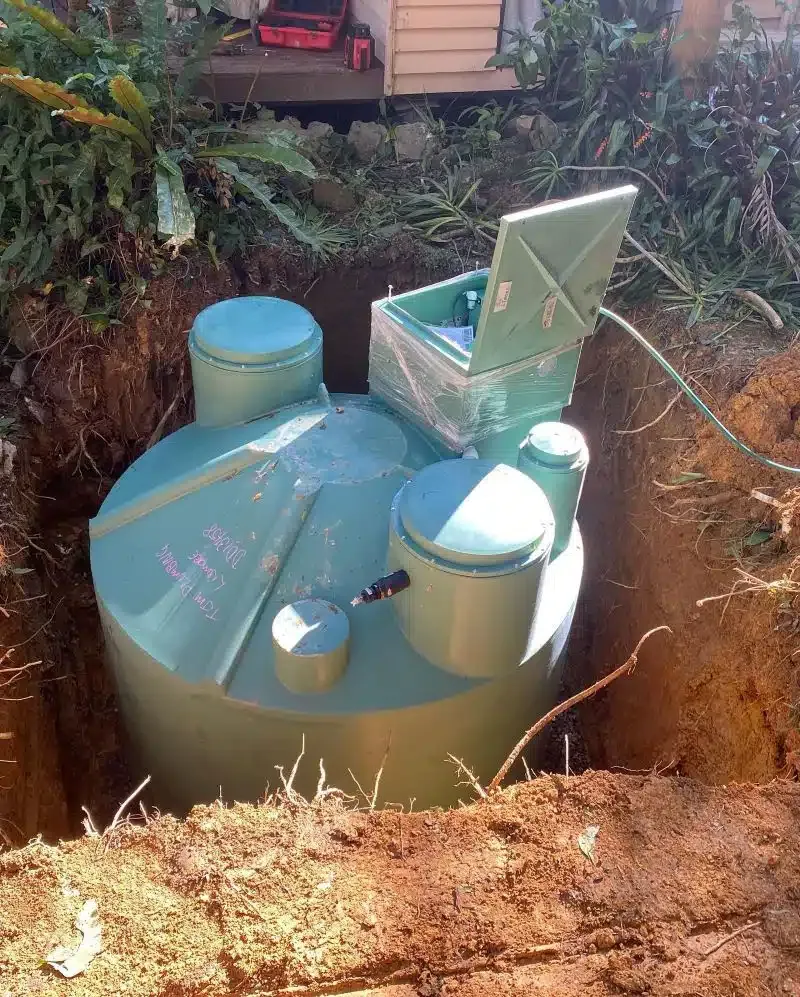 Green septic tank installed in a brown earth pit, surrounded by plants, with a house in the background.