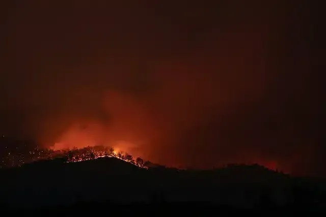 A nighttime wildfire blazes across a hillside, with orange flames and smoke against a dark sky.