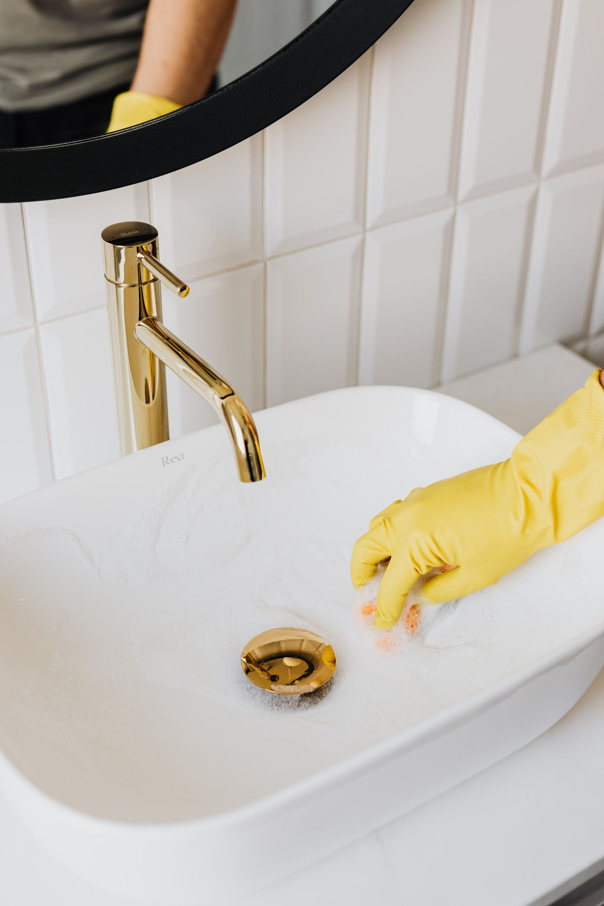 Yellow-gloved hand cleaning a white sink with a gold faucet and round mirror in a modern bathroom