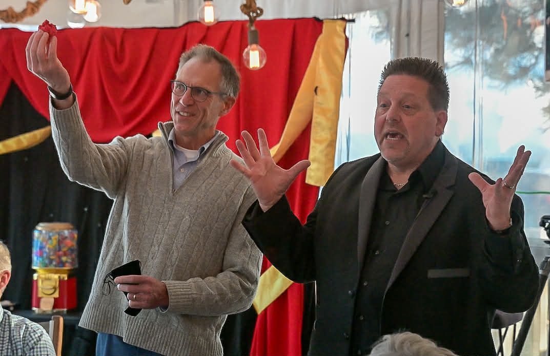 Two men performing a magic trick; one holds a small red object, the other gestures with open hands. Red curtains in the background.