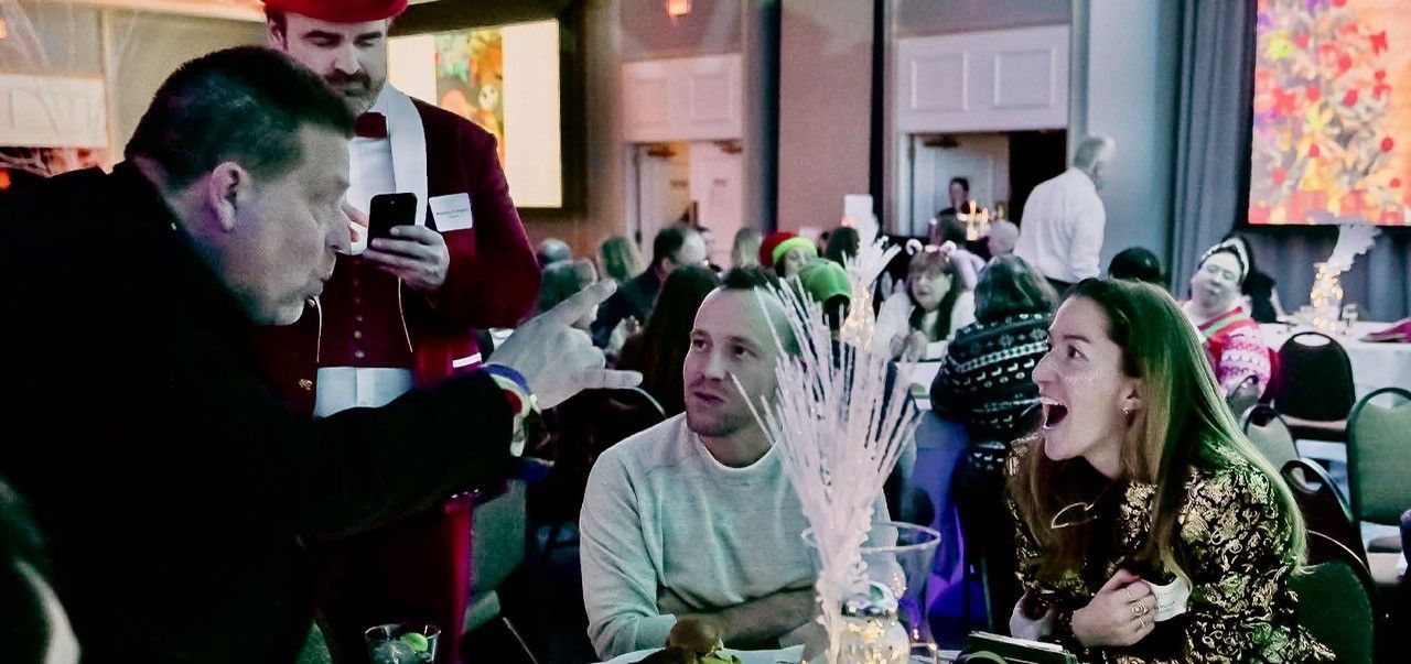 Man showing something to seated people at a festive event; Santa-like figure in background.