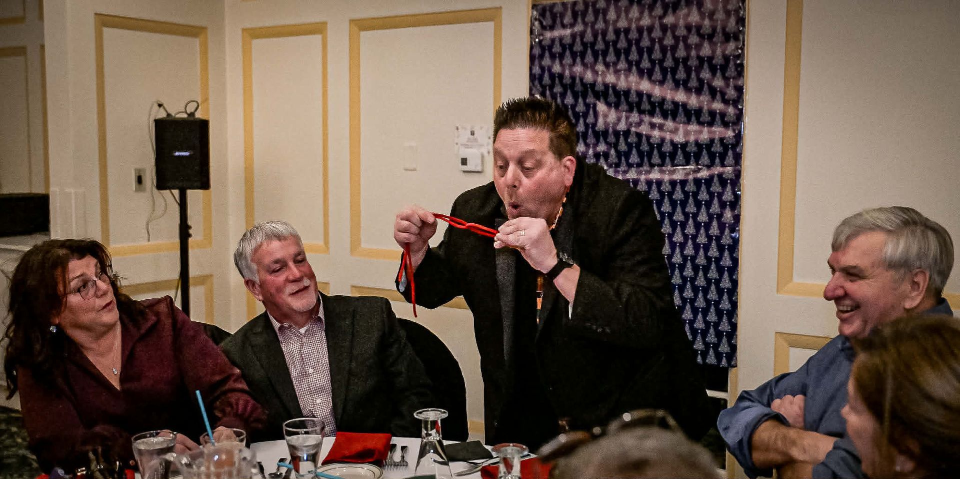 People seated at tables in a banquet hall. A man is interacting with a table of guests, who appear to be laughing.