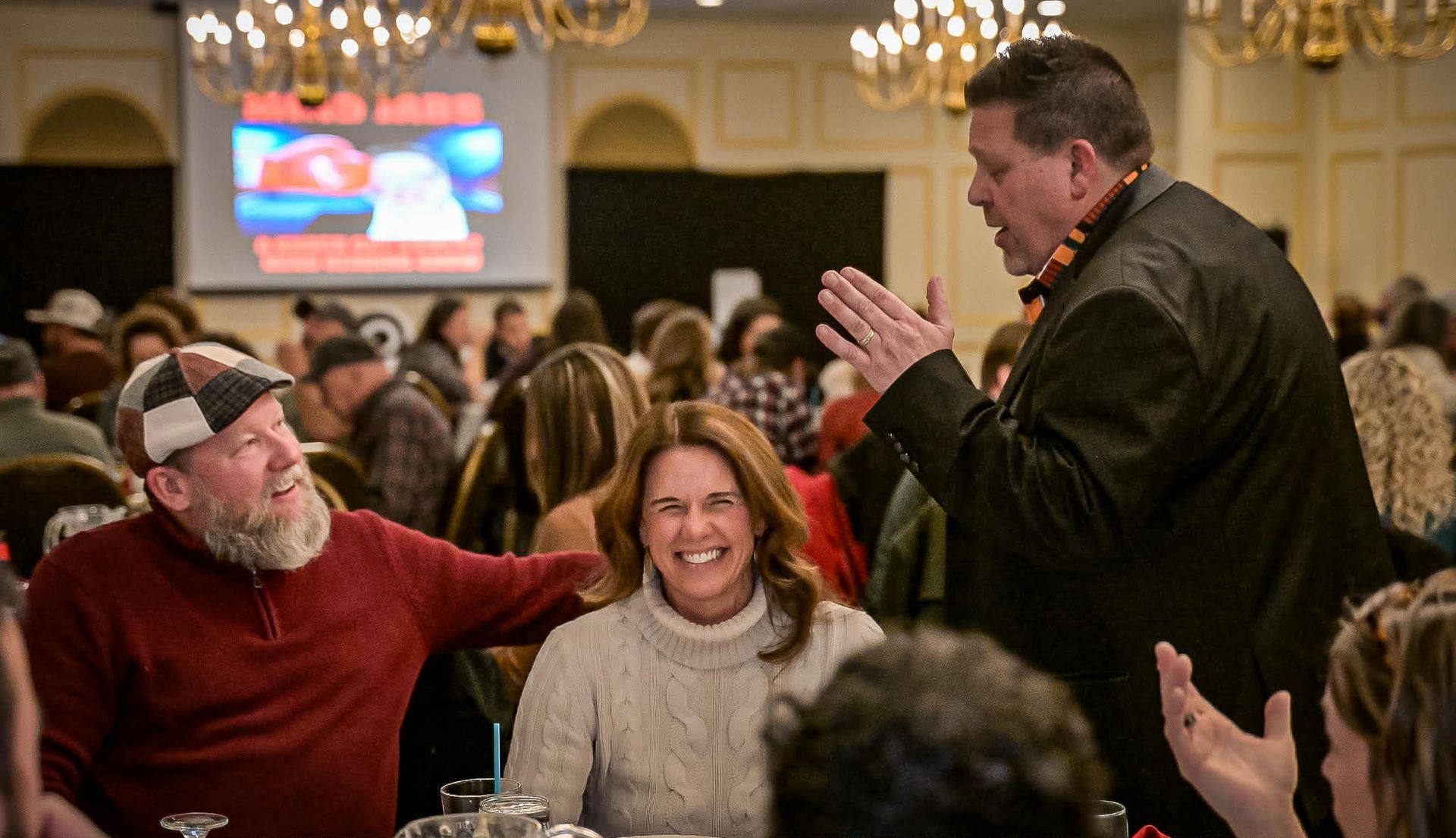 Man gesturing to two smiling people at a banquet table; a projector screen and chandeliers are in the background.
