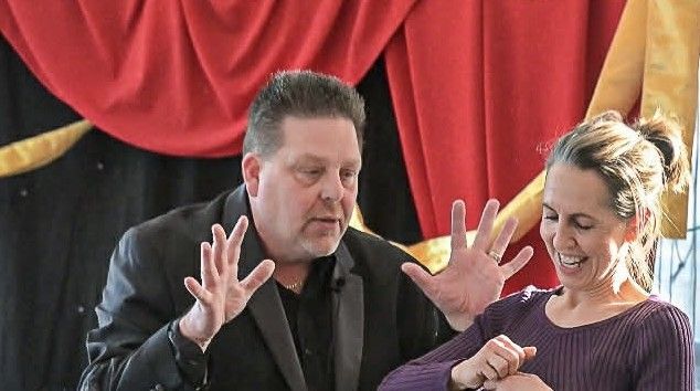 Magician gestures towards a smiling woman. Red and black curtains in background.