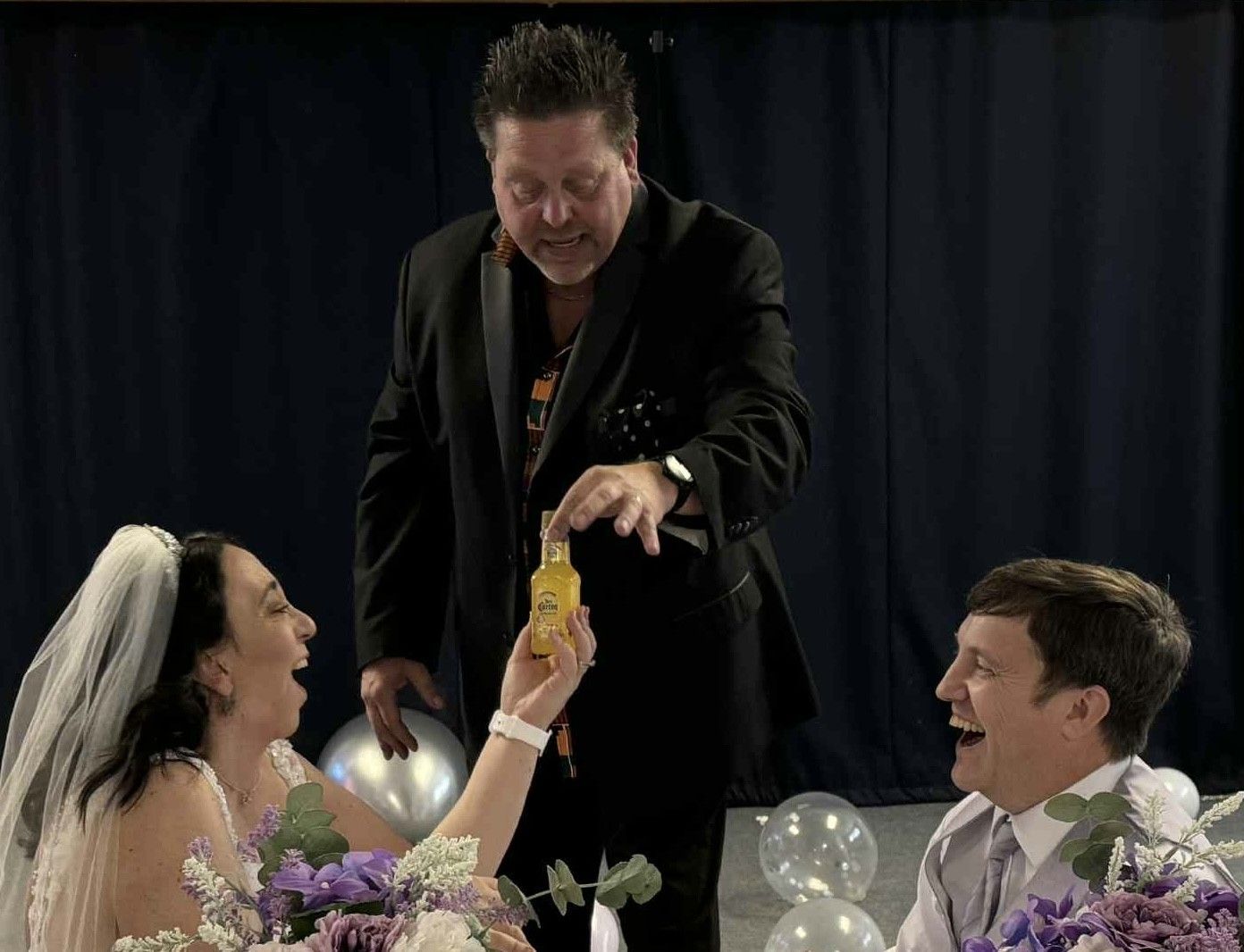 Wedding ceremony: A man offers a yellow item to a bride and groom, who are laughing.