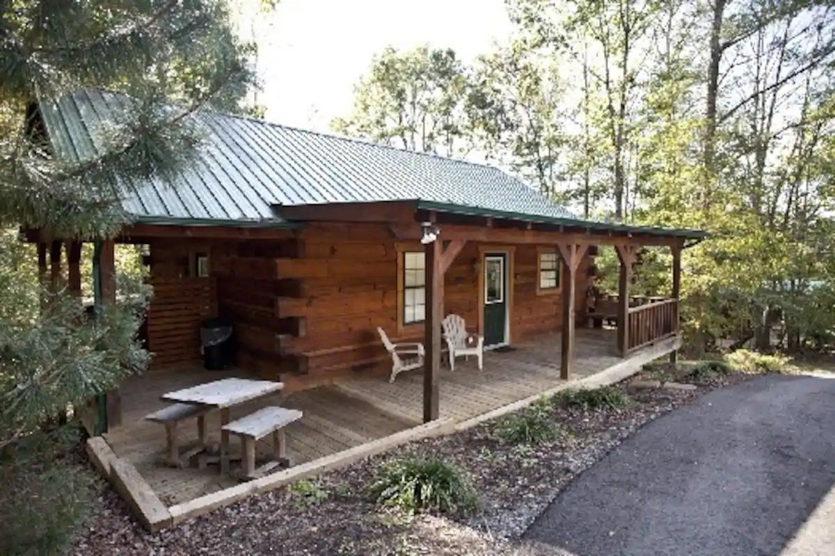 Wooden cabin with porch, picnic table, and chairs beside a paved path in a wooded area