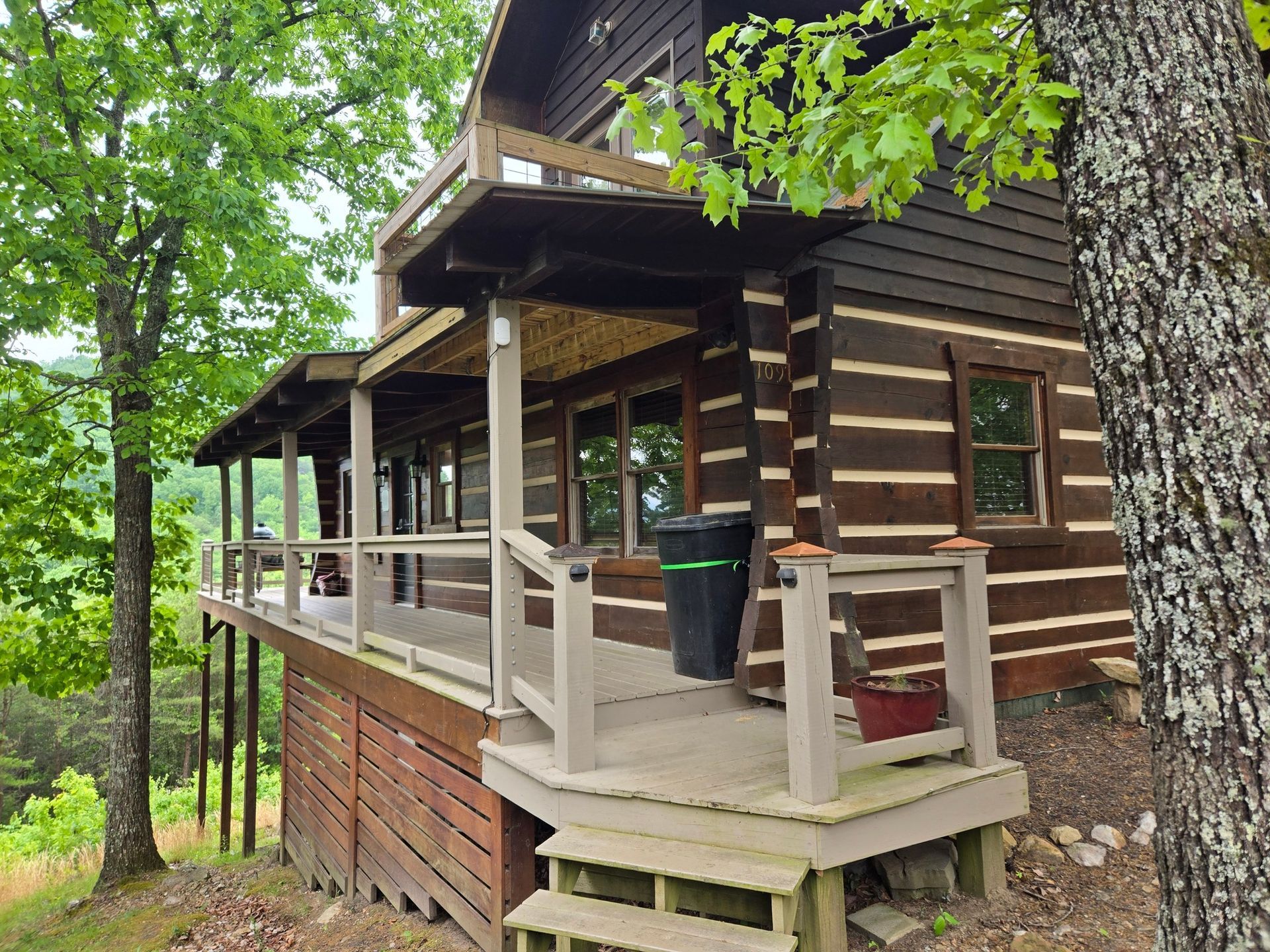 Wooden cabin porch with railing and steps, surrounded by green trees in a wooded setting