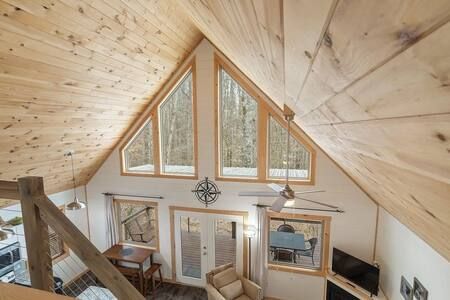 A bright loft living room with vaulted wood ceiling, large triangular windows, and cozy seating below.
