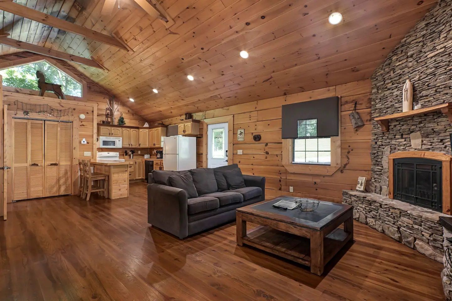 Cozy log cabin living room with vaulted wood ceiling, stone fireplace, gray sofa, and hardwood floors