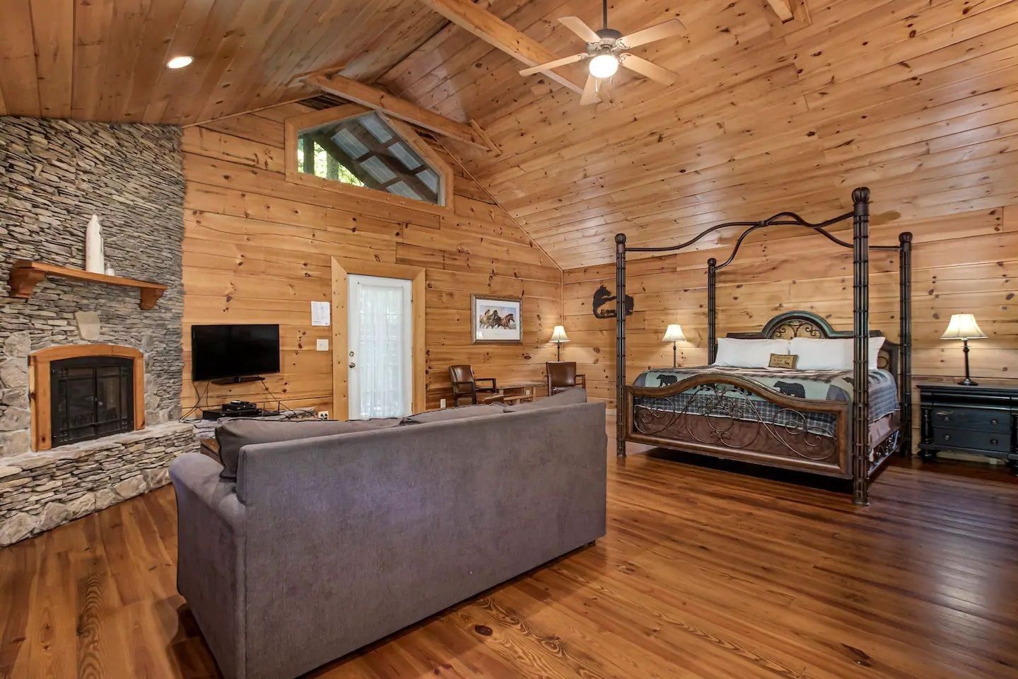 Rustic living room with stone fireplace, gray sofa, wood-paneled walls, and a four-poster bed.