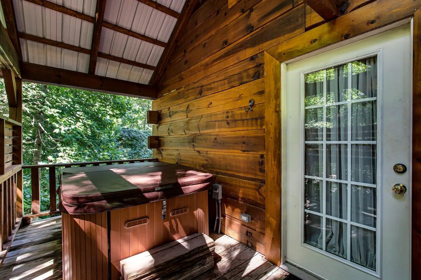Covered wooden porch with a hot tub beside a white glass-paneled door and forest view.