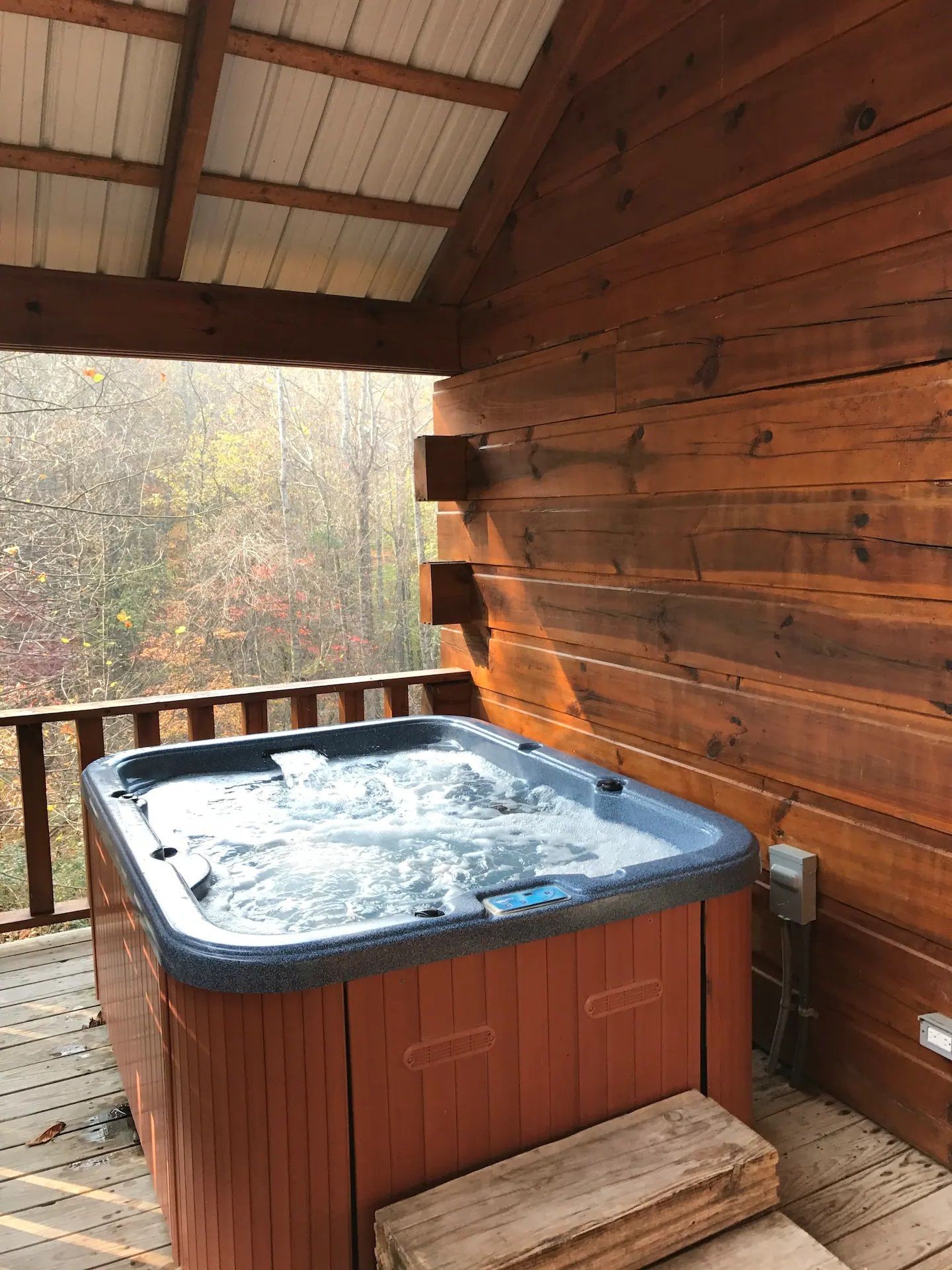 Hot tub on a wooden deck under a covered porch, beside a log wall, with a wooded view beyond.