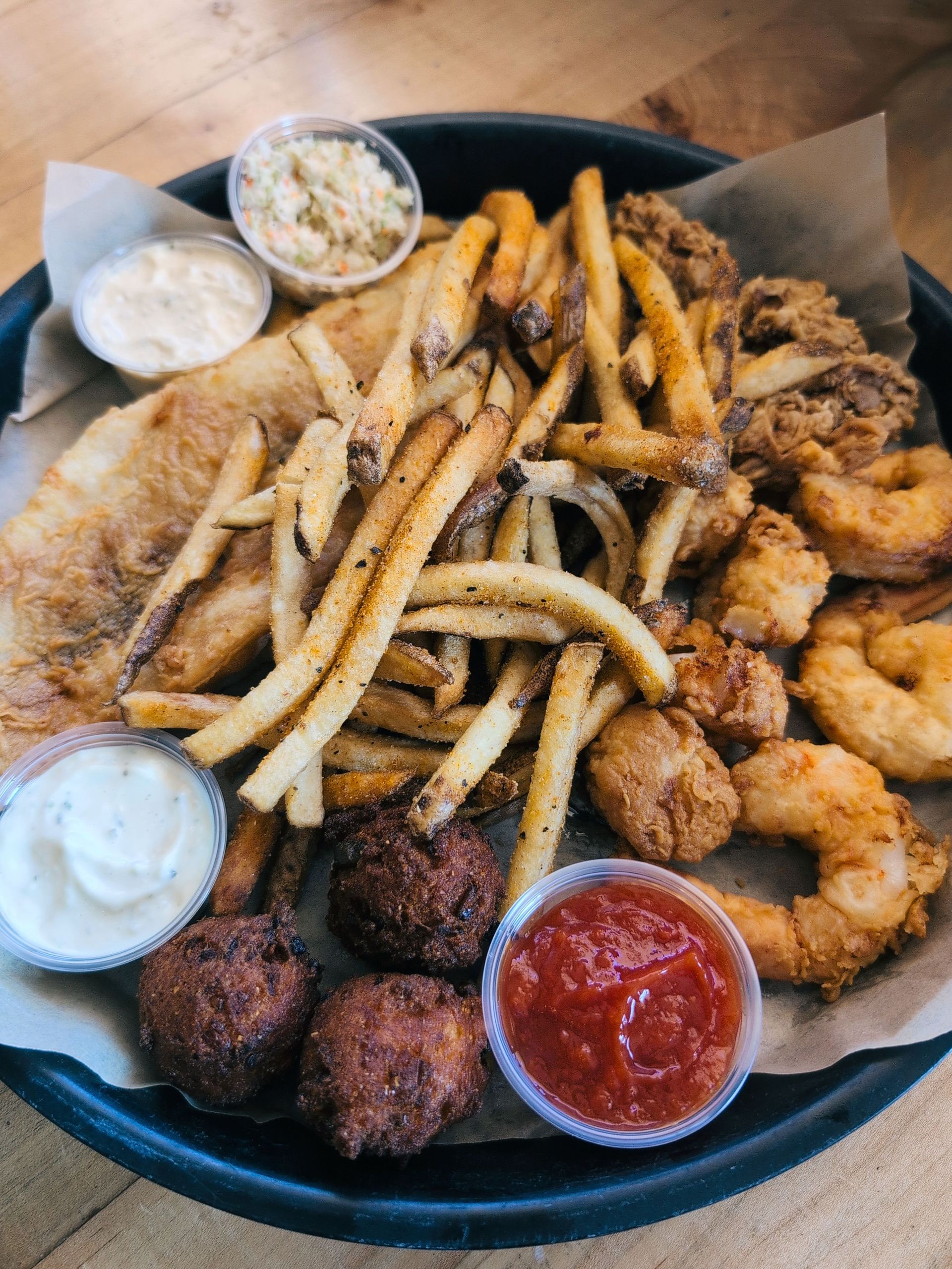 Fried seafood platter with fries, coleslaw, hush puppies, and dipping sauces on a black tray