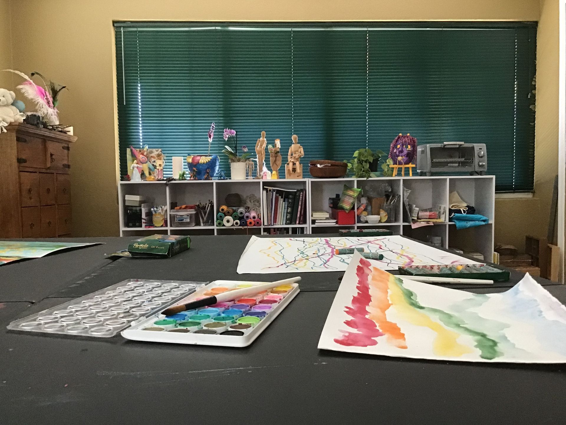 Watercolor paints and art supplies on a table with a shelf of art in the background.