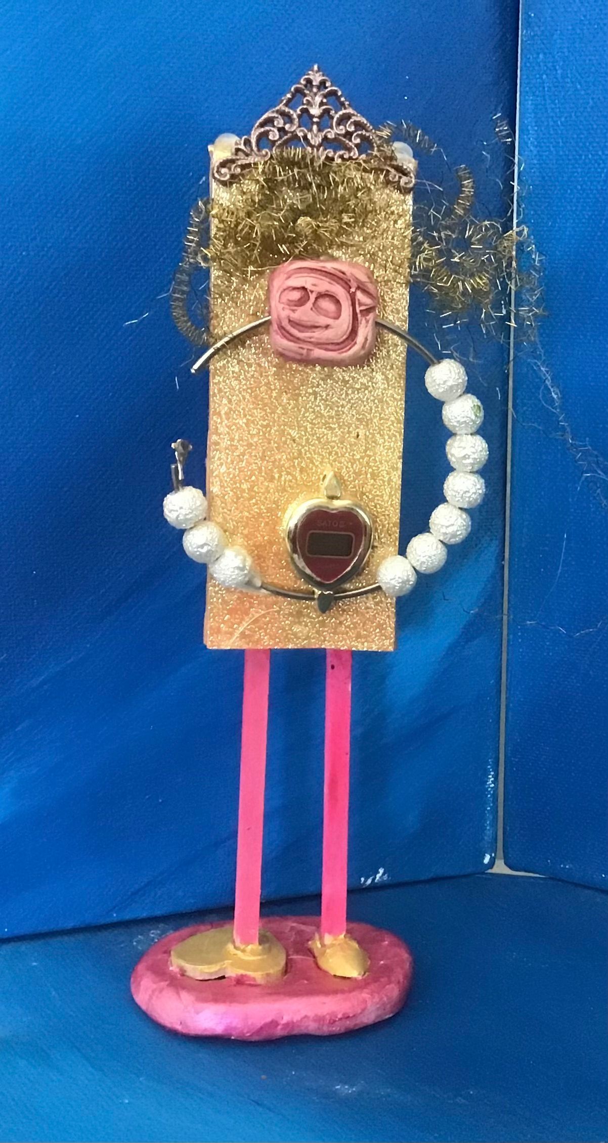 Glittery craft figure: rectangular gold torso, pink legs, gold crown, pink base, pearl bracelet.