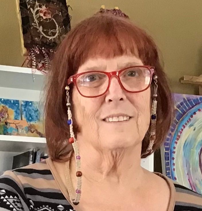 Woman with red glasses and hair, beaded eyeglass chain, in front of paintings.