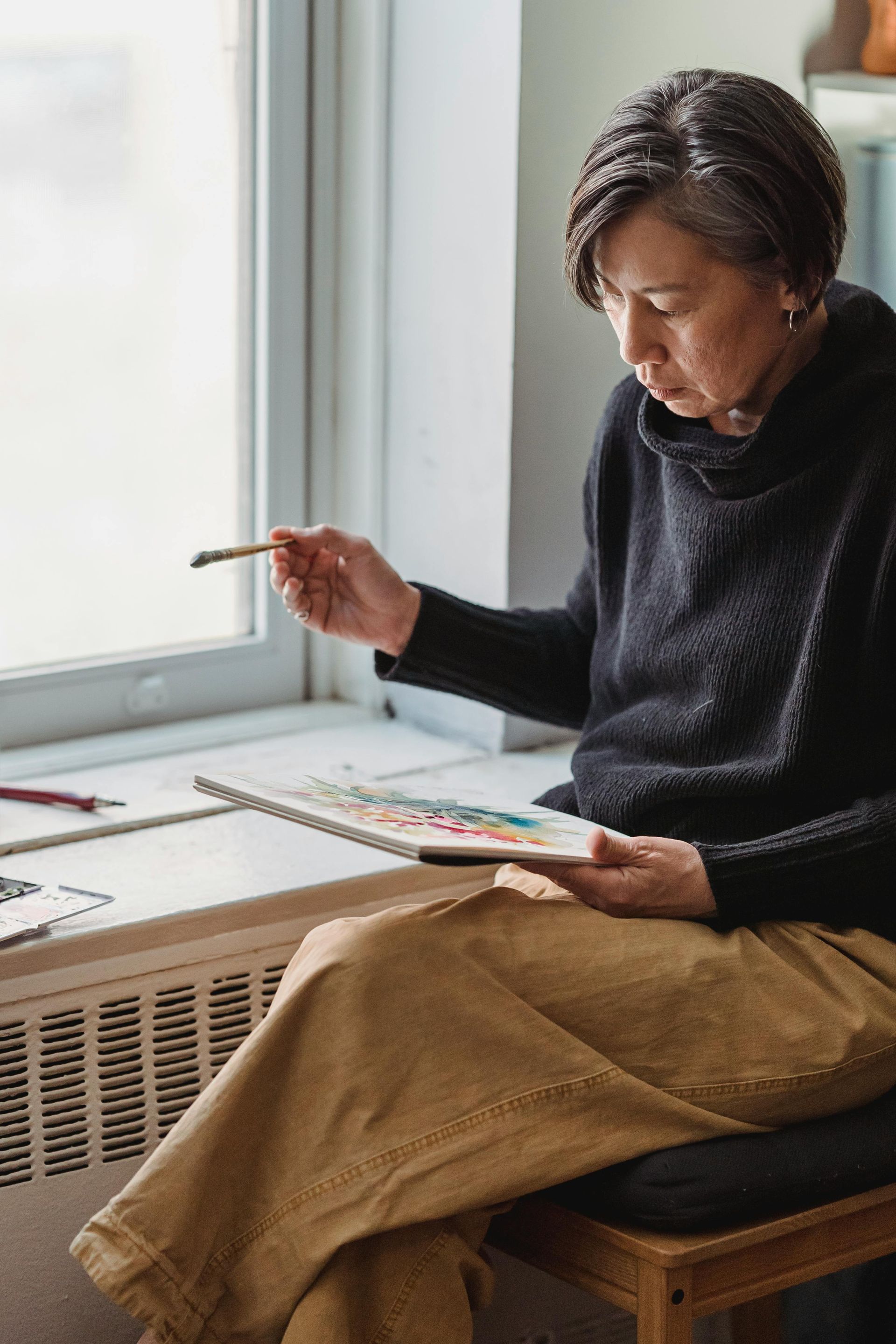 Artist seated by a window, holding a paintbrush and palette, focused on her work.