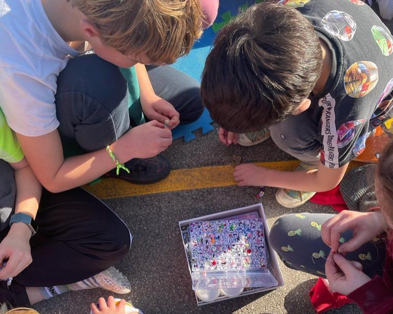 Children making colorful rubber band bracelets outdoors with a container of bands.