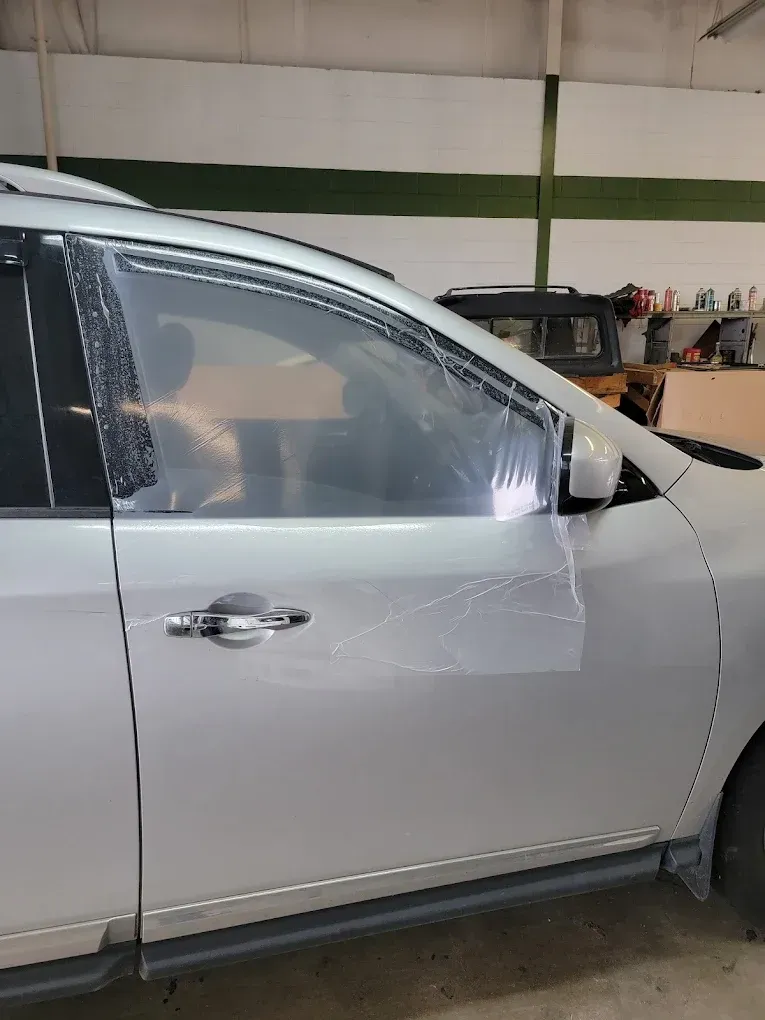 Silver car with clear protective film on the side window, likely being applied in a garage.