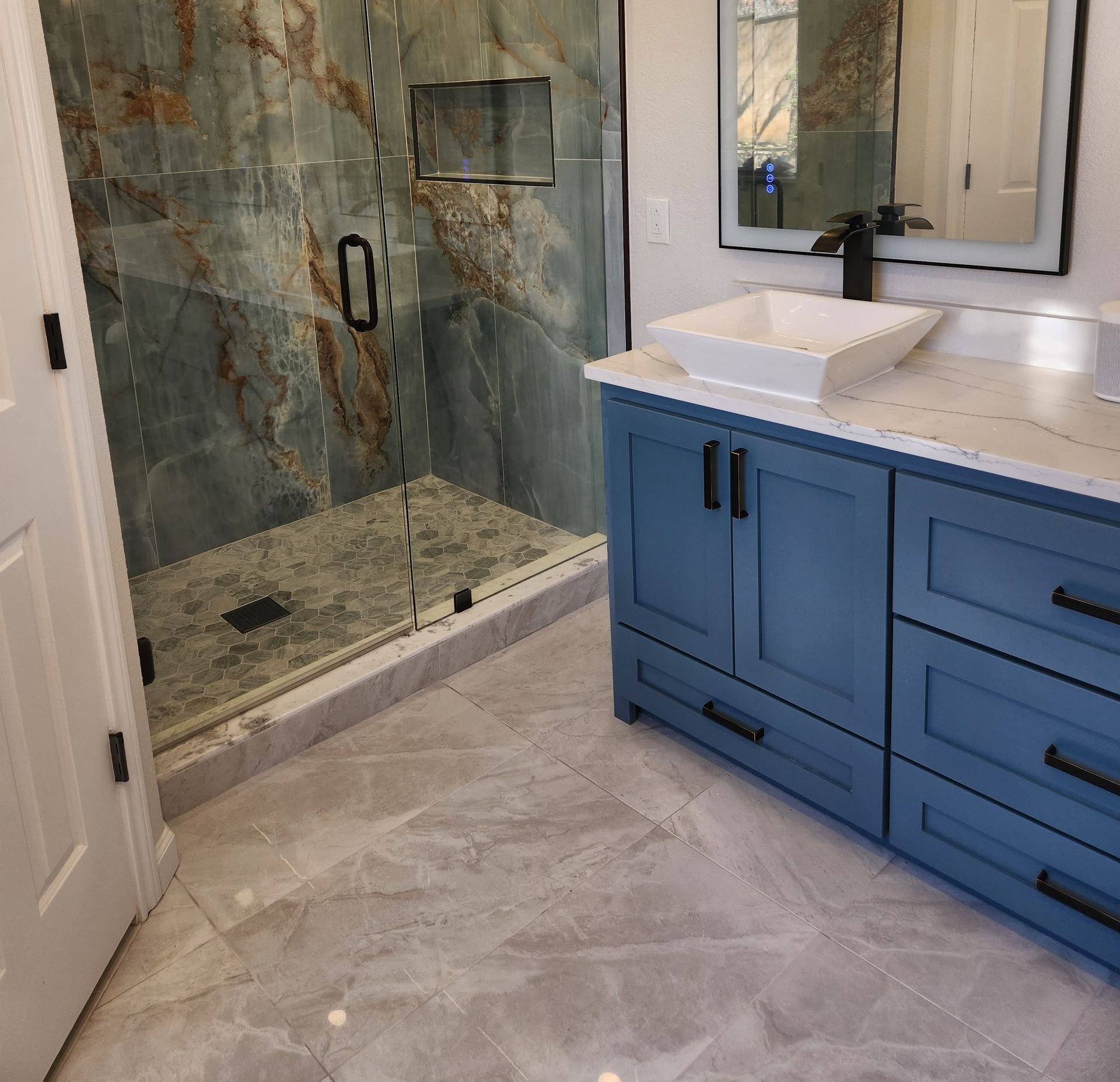 Blue bathroom vanity with white countertop and sink, next to a glass shower with marble-like wall.
