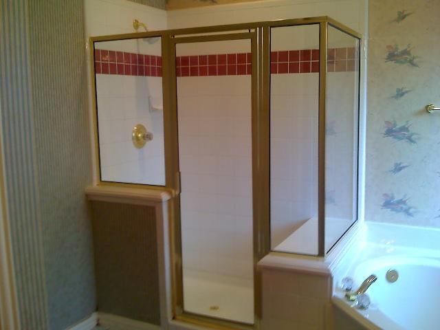 Gold-framed, glass-walled shower enclosure with red tile accents; next to a bathtub; in a bathroom with patterned wallpaper.