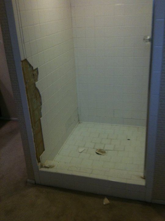 Shower stall with damaged wall; white tile and floor; visible debris.