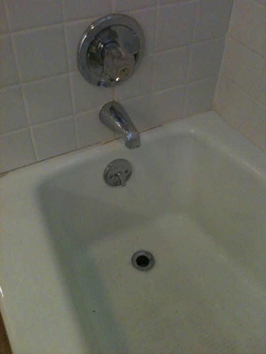 White bathtub with chrome faucet and drain, against white tiled wall.