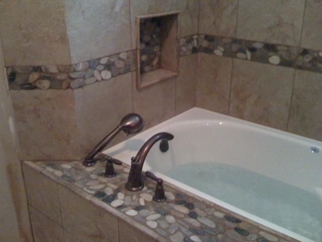 Bathroom with a white bathtub filled with water. Bronze faucet and showerhead. Pebble tile border.