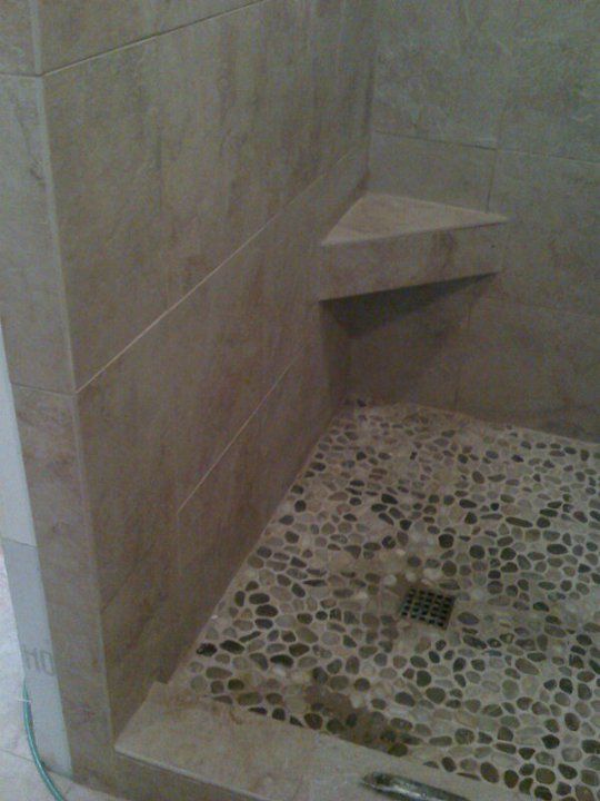 Shower with pebble tile floor and corner shelf. Light-colored wall tiles and dark drain.