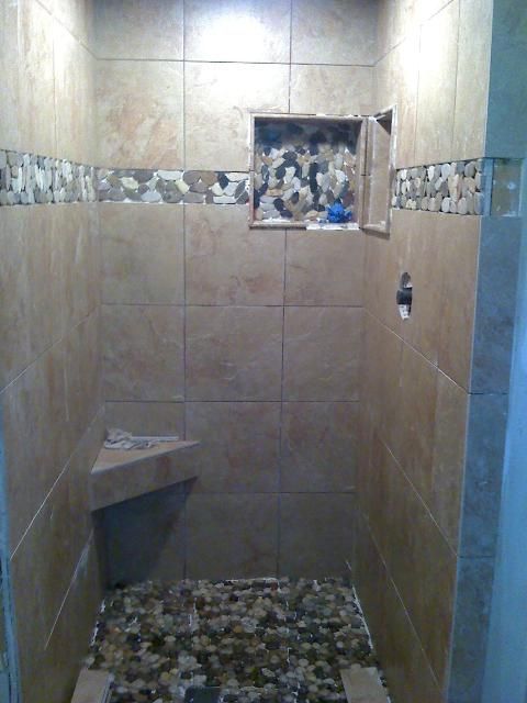 A tiled shower with a pebble floor, a built-in shelf, and a corner shelf; beige and brown tones.