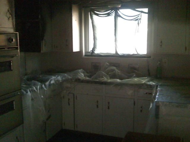 Smoke-damaged kitchen with plastic sheeting covering counters and cabinets; window with torn curtains.