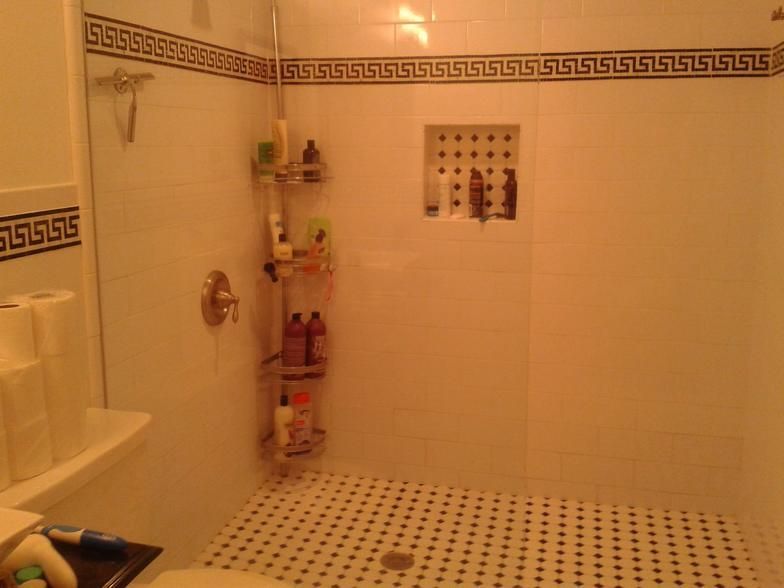 White-tiled shower with black-tiled floor, Greek key trim, built-in shelf, and a corner shower caddy holding toiletries.