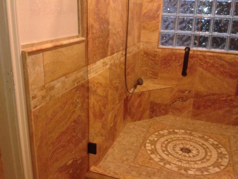 A tiled shower with a patterned floor. Brown and tan tiles, glass block window, black handle.