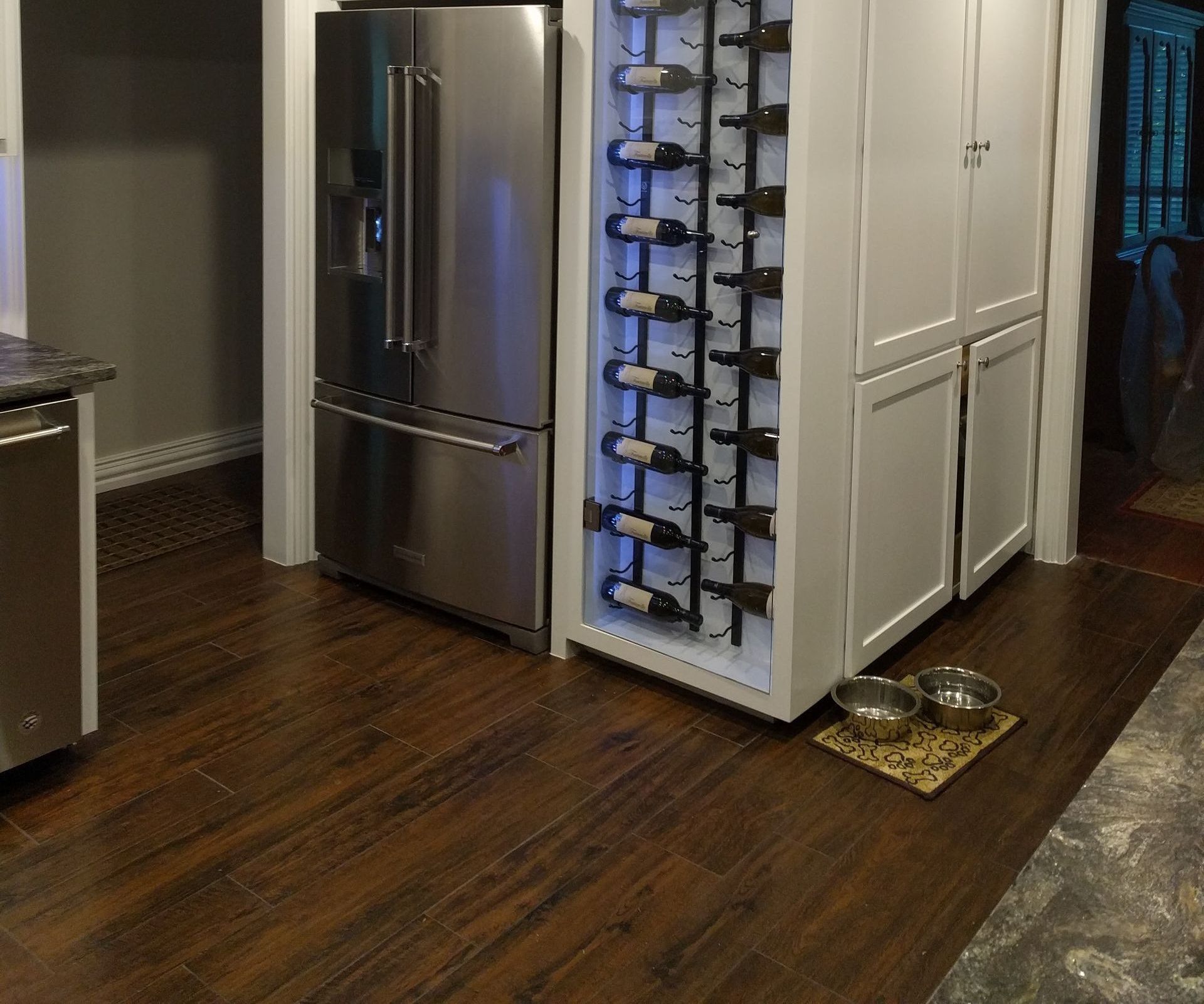 Stainless steel refrigerator next to a wine rack, and white cabinets. Brown flooring.
