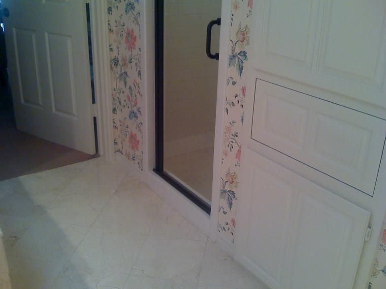 Bathroom with white tile floor, glass shower, white cabinets, and floral wallpaper.