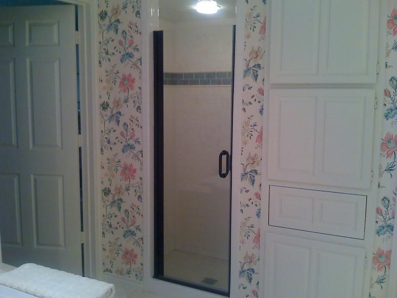 Bathroom with a glass shower, white cabinets, floral wallpaper, and a closed white door.