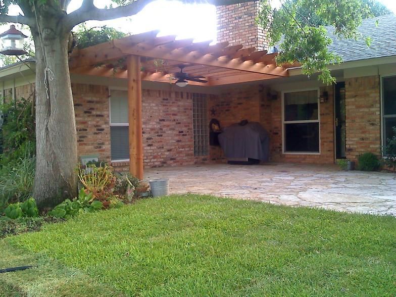 Wooden pergola over a brick patio, attached to a house with a green lawn and tree in the foreground.