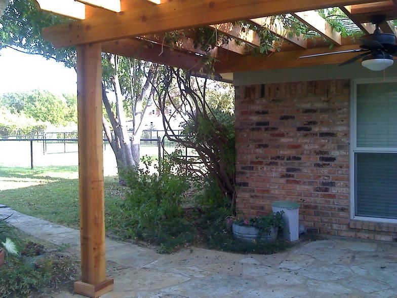 Wooden pergola over stone patio next to brick wall and bushes.