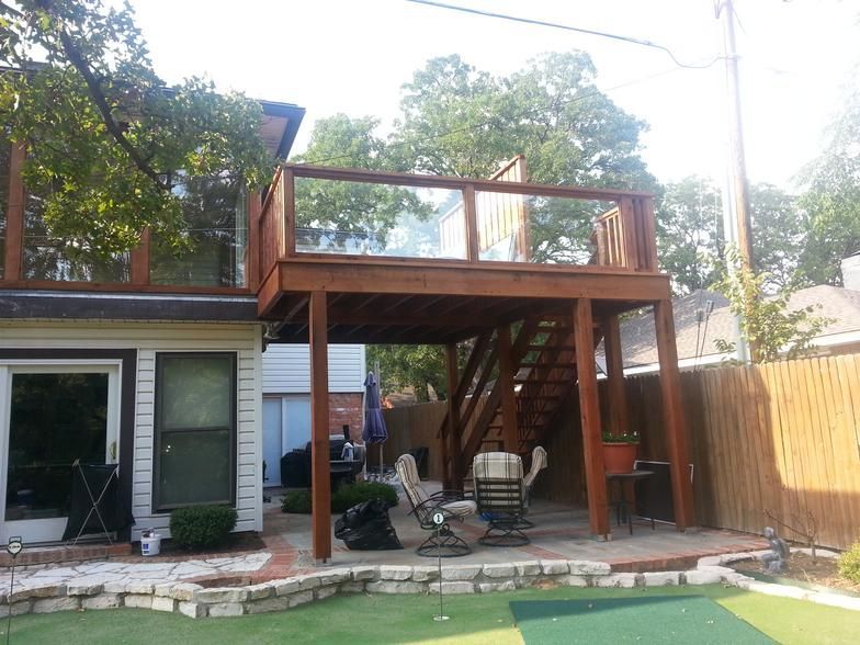 Two-story wooden deck with glass railing, stairs, and patio below. Backyard setting, partially shaded.
