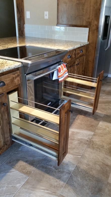 Open kitchen cabinet with pull-out spice racks flanking a stainless steel oven. Dark wood cabinets and granite countertops.