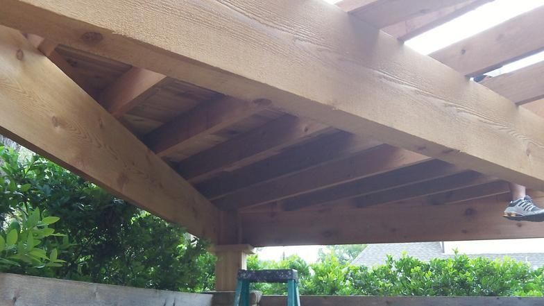 Wooden pergola roof with angled support beams, partially shaded, outdoor setting.