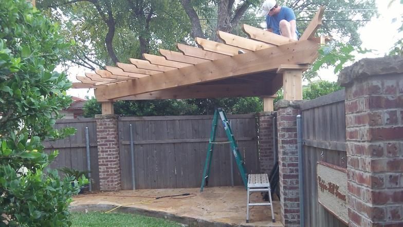Man on a wooden pergola roof over brick pillars, working on construction. Green ladder present.