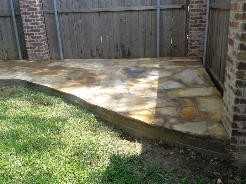 Stone patio with a concrete border, adjacent to grass and a wooden fence.