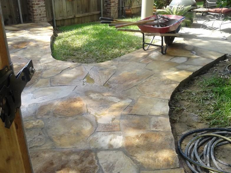 Stone pathway leading from a gate, bordering grass and a wheelbarrow in a sunny outdoor setting.