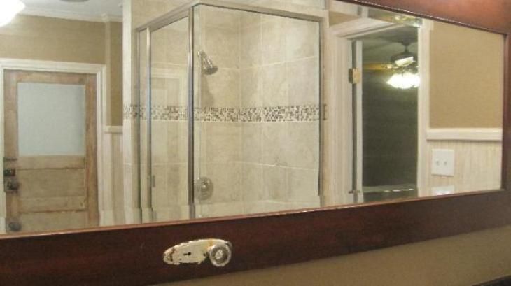 Mirror reflecting a bathroom with a shower, door, and a light switch. Brown and beige tones.