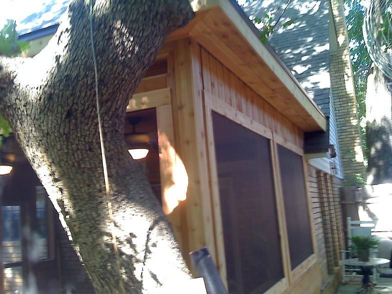 Wooden screened-in porch next to a tree. Brown wood, dark screens, and a light-colored roof.