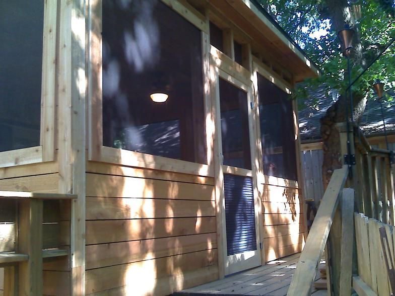 Wooden screened-in porch with windows and a light fixture, built around a tree, on a sunny day.