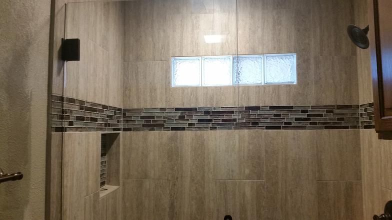 Shower with beige and brown tiled walls, glass door, dark hardware, and decorative mosaic band.